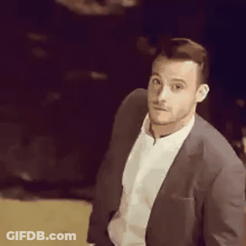 A Man In A Suit And White Shirt Is Standing In A Dark Room Looking At The Camera . GIF