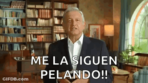 A Man In A Suit And White Shirt Is Standing In A Living Room With A Bookshelf In The Background . GIF