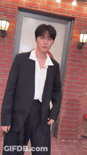 A Man In A Suit And White Shirt Is Standing In Front Of A Red Brick Wall . GIF