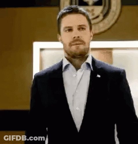 A Man In A Suit And White Shirt Is Standing In Front Of A Wall With A Seal On It . GIF
