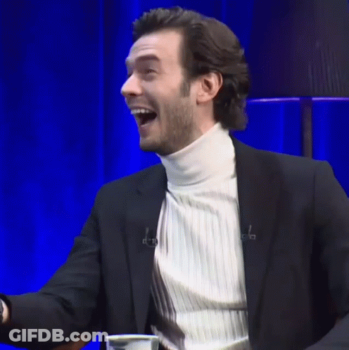 A Man In A Suit And White Turtleneck Is Laughing With His Mouth Open GIF