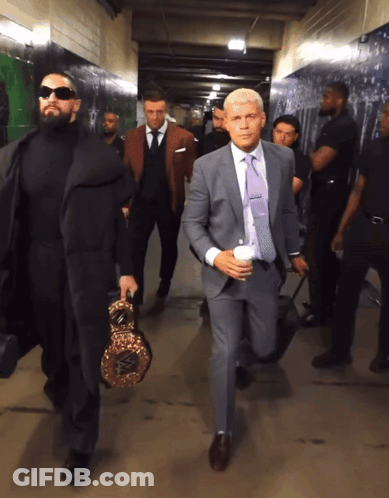 A Man In A Suit Carrying A Boxing Ring Is Walking Down A Hallway GIF