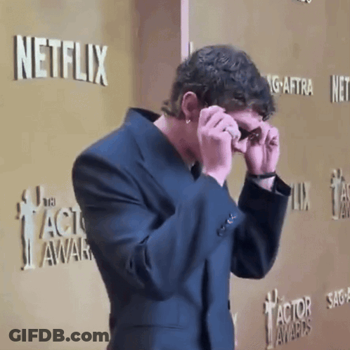 A Man In A Suit Covering His Eyes In Front Of A Netflix Sign GIF