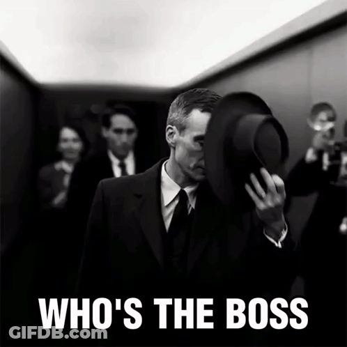 A Man In A Suit Covering His Face With His Hat And The Words Who 'S The Boss Behind Him GIF