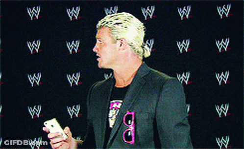 A Man In A Suit Holds A Cell Phone In Front Of A Wall With Wrestlers Logos On It GIF