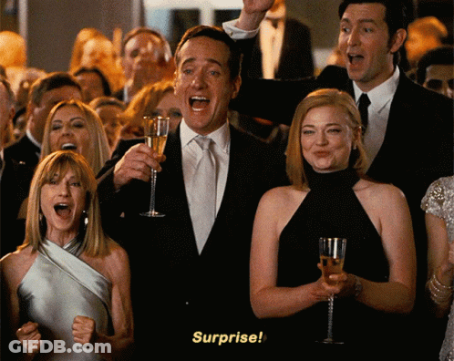 A Man In A Suit Holds A Glass Of Champagne In Front Of A Crowd That Says Surprise GIF