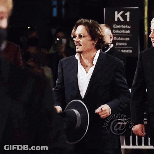 A Man In A Suit Holds A Hat Over His Face In Front Of A Sign That Says K1 GIF