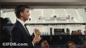 A Man In A Suit Holds A Hot Dog In Front Of A Stadium GIF