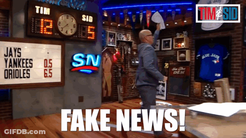 A Man In A Suit Holds A Piece Of Paper In Front Of A Scoreboard That Says Fake News GIF