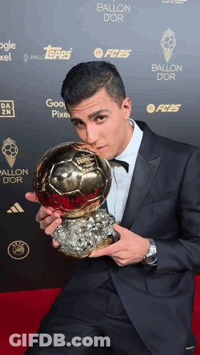 A Man In A Suit Holds A Soccer Ball In Front Of A Wall That Says Ballon Dor GIF