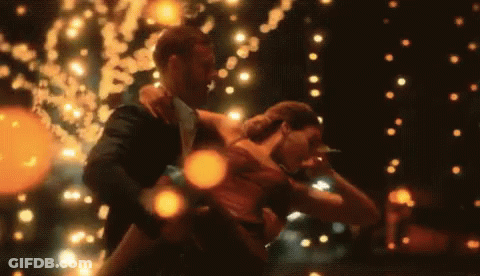 A Man In A Suit Holds A Woman In His Arms While They Dance In Front Of A String Of Lights GIF