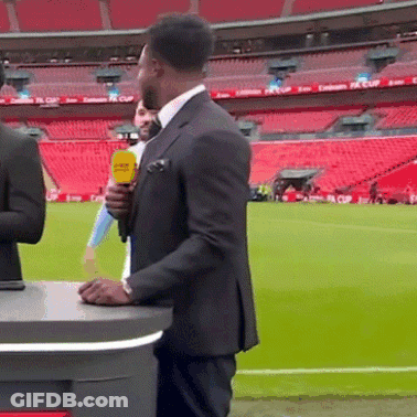 A Man In A Suit Holds A Yellow Microphone In Front Of A Stadium That Has Emirates Fa Cup Written On The Seats GIF