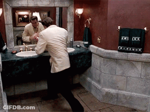 A Man In A Suit Is Brushing His Teeth In Front Of A Bathroom Mirror GIF