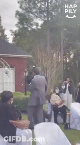 A Man In A Suit Is Carrying A Baby In His Arms While A Group Of People Are Sitting In Chairs . GIF