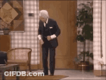 A Man In A Suit Is Dancing In A Living Room With A Chair And A Plant . GIF