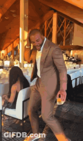 A Man In A Suit Is Dancing In A Restaurant With A Woman Sitting In A Chair Behind Him GIF