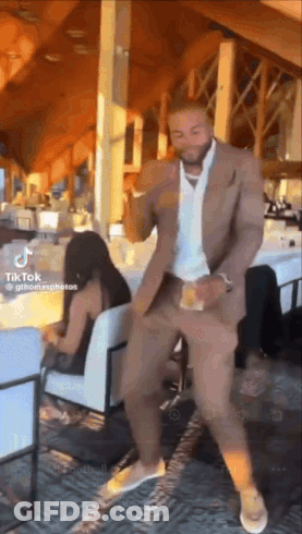 A Man In A Suit Is Dancing In A Restaurant With A Woman Sitting In A Chair Behind Him . GIF