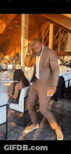 A Man In A Suit Is Dancing In A Room With A Woman Sitting In A Chair GIF