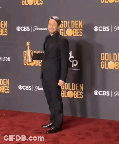 A Man In A Suit Is Dancing On A Red Carpet In Front Of A Wall That Says Golden Globes GIF