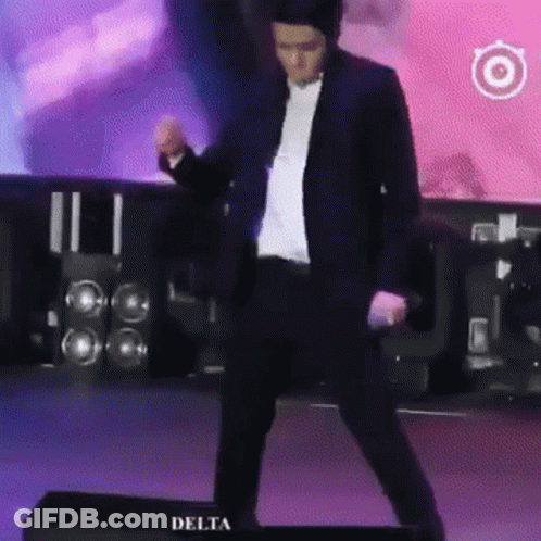 A Man In A Suit Is Dancing On A Stage In Front Of Delta Speakers GIF