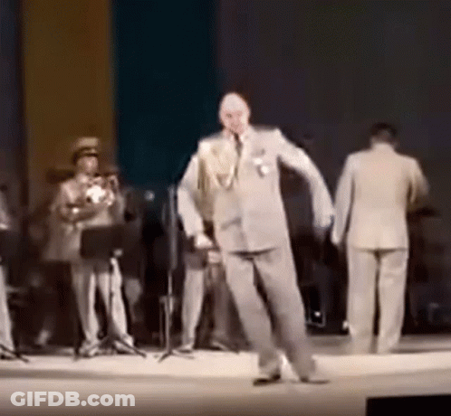 A Man In A Suit Is Dancing On A Stage While A Band Plays Behind Him . GIF