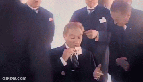 A Man In A Suit Is Drinking A Cup Of Coffee And Smoking A Cigarette While A Group Of Men Look On . GIF