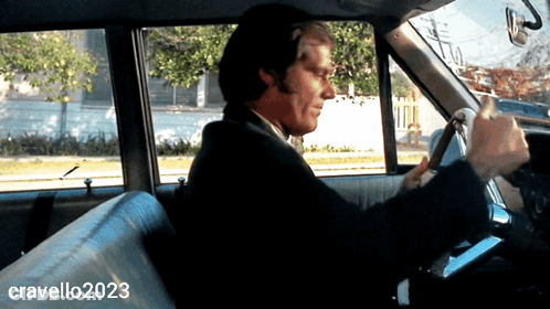 A Man In A Suit Is Driving A Car With Cravello2023 Written On The Bottom Right GIF