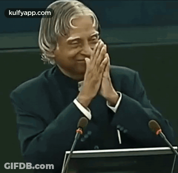A Man In A Suit Is Giving A Speech With His Hands Folded In Front Of Two Microphones . GIF