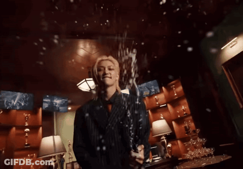 A Man In A Suit Is Holding A Bottle Of Champagne In A Room Filled With Trophies GIF