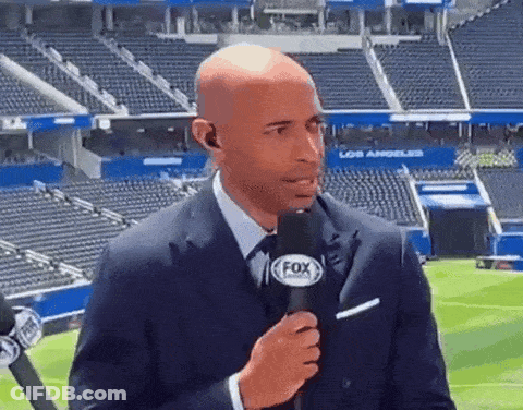 A Man In A Suit Is Holding A Fox Microphone In Front Of A Stadium . GIF