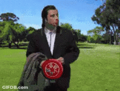 A Man In A Suit Is Holding A Frisbee In A Park With Trees In The Background GIF