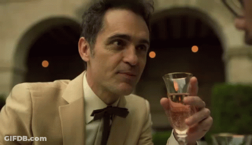 A Man In A Suit Is Holding A Glass Of Wine In His Hand . GIF