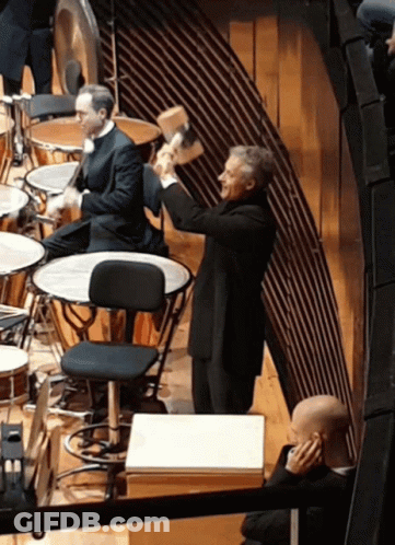 A Man In A Suit Is Holding A Hammer In His Hand In Front Of A Drum Set GIF