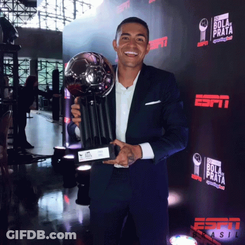 A Man In A Suit Is Holding A Trophy In Front Of A Wall That Says Bola De Prata GIF