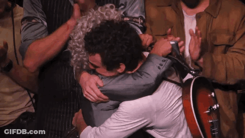 A Man In A Suit Is Hugging Another Man While Holding A Guitar GIF