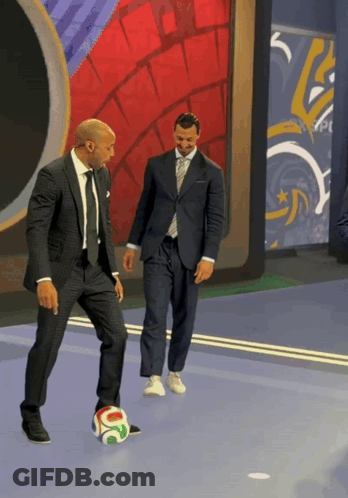 A Man In A Suit Is Kicking A Soccer Ball While Another Man Watches GIF