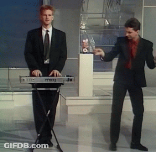 A Man In A Suit Is Playing A Keyboard While Another Man Dances Behind Him GIF