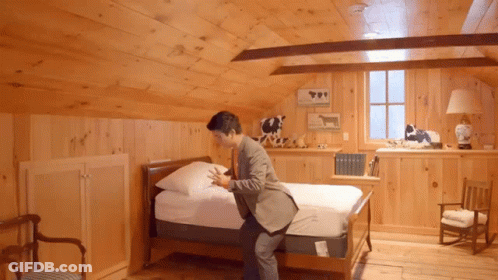 A Man In A Suit Is Putting A Mattress On A Bed In A Wooden Room GIF