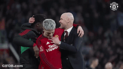 A Man In A Suit Is Putting His Hand On A Man In A Red Teamviewer Jersey GIF