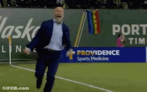 A Man In A Suit Is Running On A Soccer Field In Front Of A Providence Sports Medicine Banner . GIF