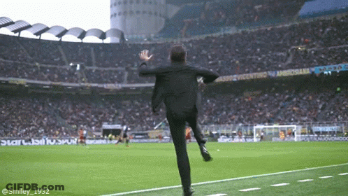 A Man In A Suit Is Running On A Soccer Field In Front Of A Stadium That Says Ambrosiaba GIF