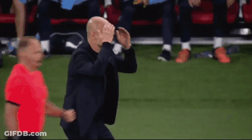 A Man In A Suit Is Running On A Soccer Field With A Referee In A Red Shirt Behind Him GIF