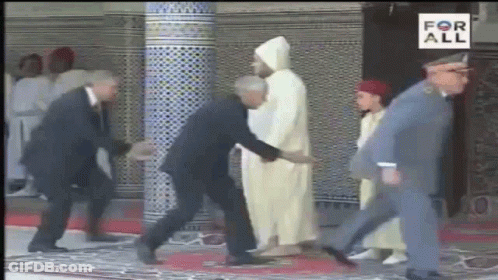 A Man In A Suit Is Shaking Hands With A Man In A White Robe While Walking Down A Red Carpet . GIF