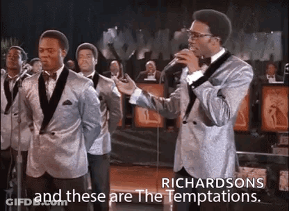 A Man In A Suit Is Singing Into A Microphone In Front Of A Group Of Men In Suits . GIF