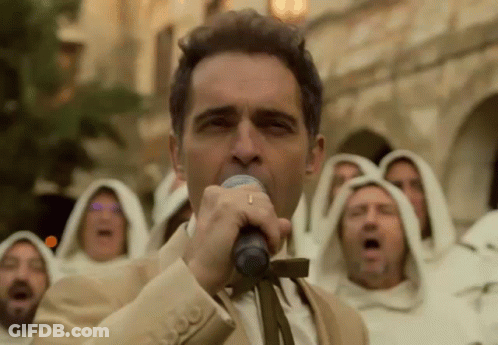A Man In A Suit Is Singing Into A Microphone In Front Of A Group Of Monks In White Robes . GIF