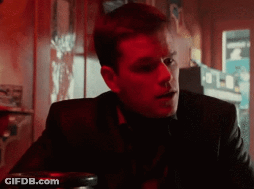 A Man In A Suit Is Sitting At A Table In A Dark Room With A Red Light Behind Him . GIF