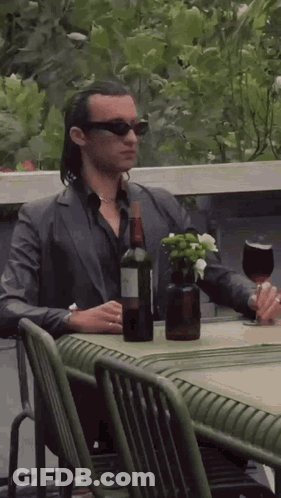 A Man In A Suit Is Sitting At A Table With A Bottle Of Wine And A Glass . GIF