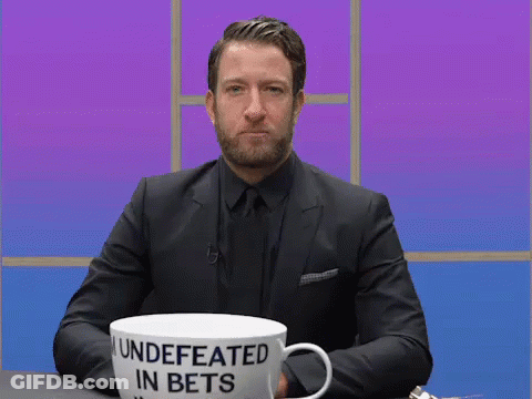 A Man In A Suit Is Sitting At A Table With A Cup That Says " Undefeated In Bets " GIF