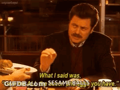 A Man In A Suit Is Sitting At A Table With A Plate Of Food And Talking To Another Man . GIF