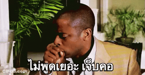 A Man In A Suit Is Sitting At A Table With His Hand In His Mouth And A Plant In The Background . GIF
